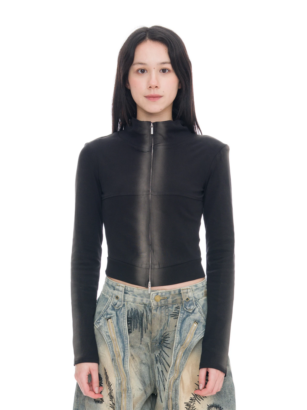 A model stands against a seamless white studio background wearing a fitted black long-sleeve top with a high collar and a full-length front zipper, paired with extremely baggy wide-leg pants in a beige, blue, and gray abstract landscape print, and black shoes.