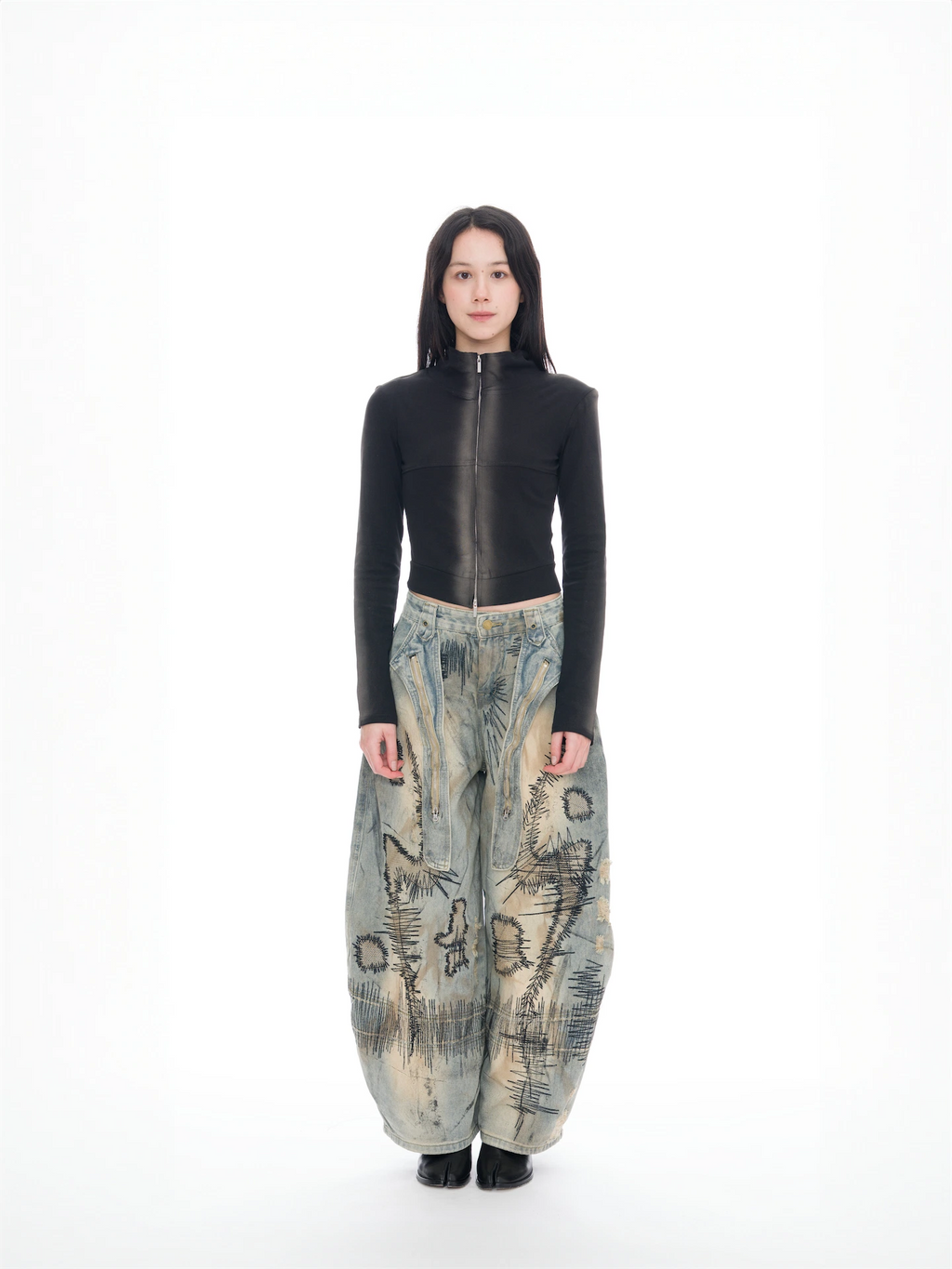 A model stands against a seamless white studio background wearing a fitted black long-sleeve top with a high collar and a full-length front zipper, paired with extremely baggy wide-leg pants in a beige, blue, and gray abstract landscape print, and black shoes.