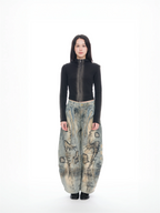 A model stands against a seamless white studio background wearing a fitted black long-sleeve top with a high collar and a full-length front zipper, paired with extremely baggy wide-leg pants in a beige, blue, and gray abstract landscape print, and black shoes.
