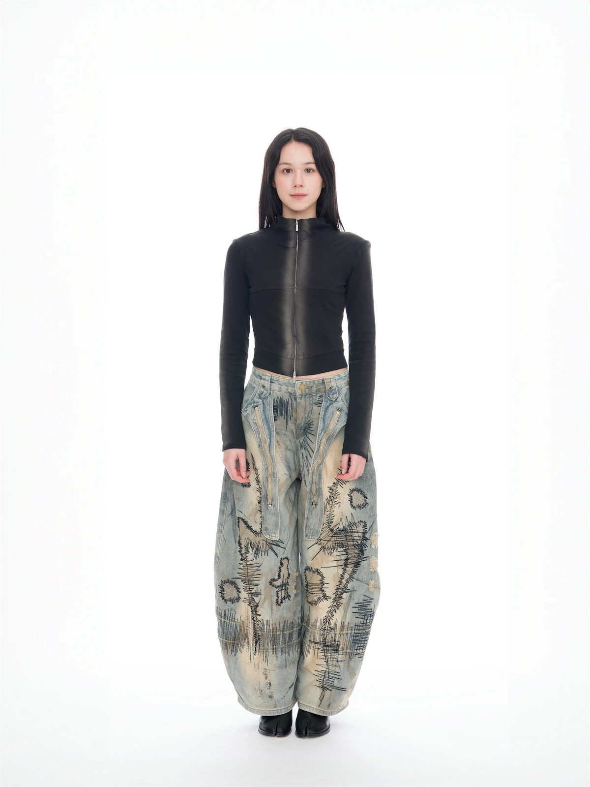 A model stands against a seamless white studio background wearing a fitted black long-sleeve top with a high collar and a full-length front zipper, paired with extremely baggy wide-leg pants in a beige, blue, and gray abstract landscape print, and black shoes.