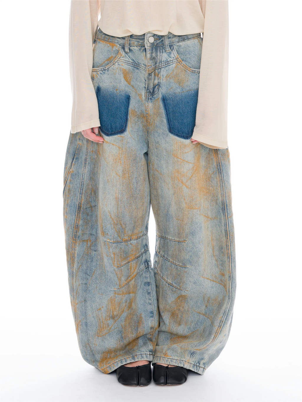 A person wearing very wide-leg light-wash denim jeans with large blue patch pockets and a distressed tan wash, paired with a cream long-sleeve top and black shoes against a white studio background.
