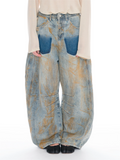 A person wearing very wide-leg light-wash denim jeans with large blue patch pockets and a distressed tan wash, paired with a cream long-sleeve top and black shoes against a white studio background.