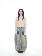 A model stands against a seamless white studio background wearing a light beige long-sleeve knit top and very wide-leg, baggy light-wash denim pants with blue patch pockets and a subtle gold-toned abstract pattern.