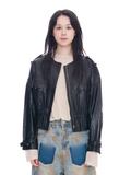 A model wearing a black leather jacket over a cream long-sleeve top and blue denim jeans with a yellow paint-splatter pattern, shot against a solid white background.