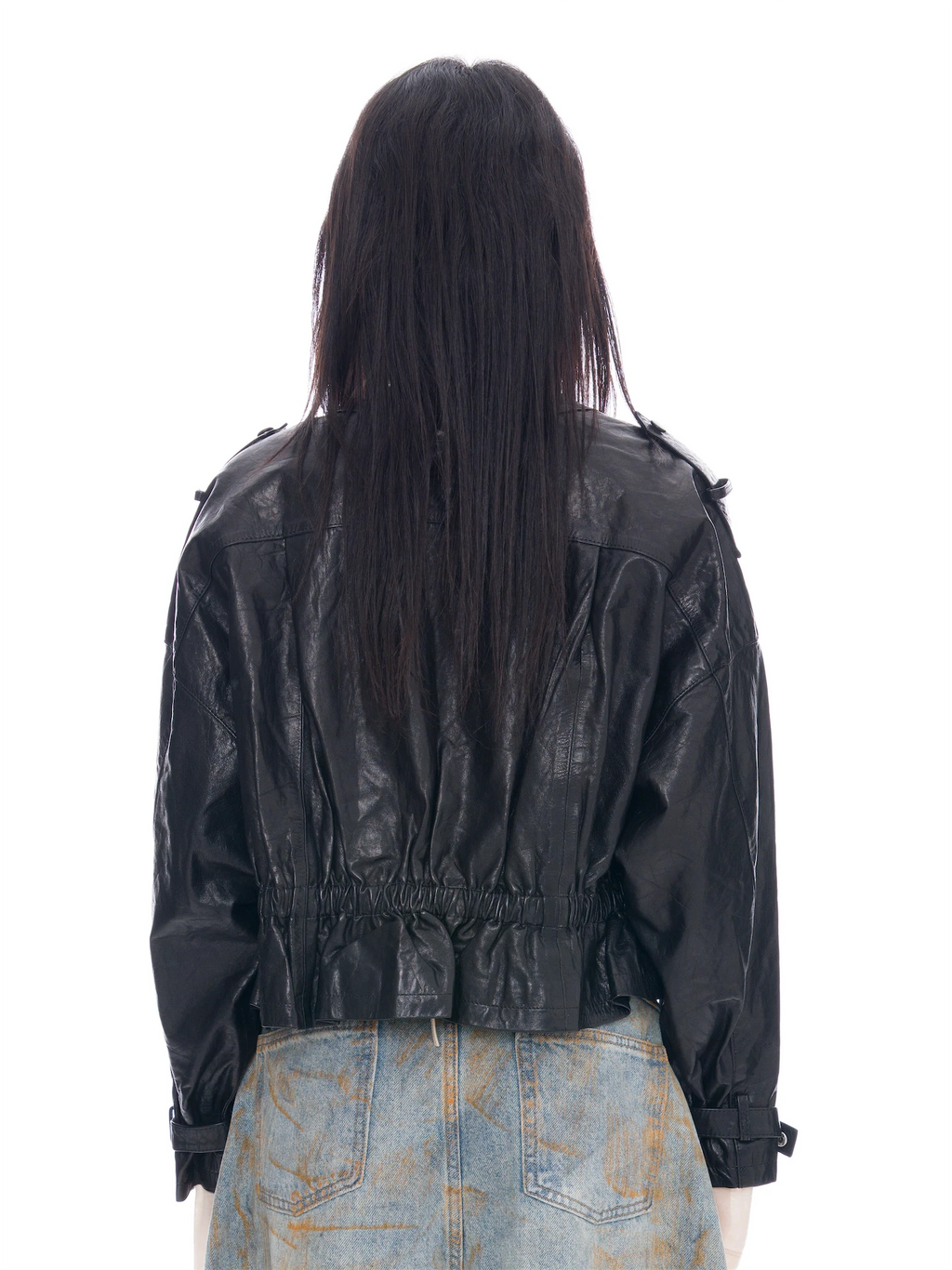 Back view of a model with long dark hair wearing a black leather jacket and light blue denim jeans with orange paint splatters, shot against a solid white background.