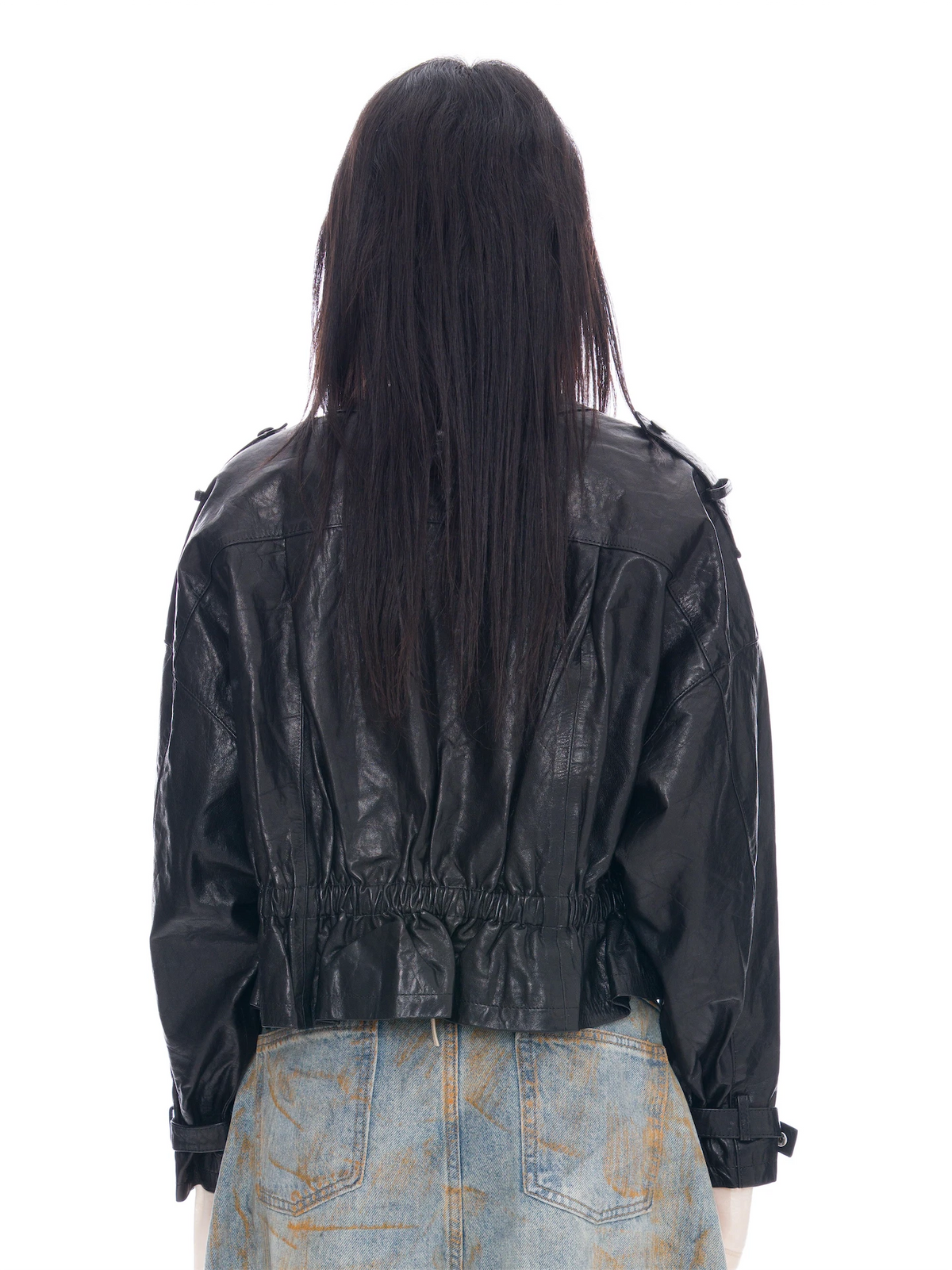 Back view of a model with long dark hair wearing a black leather jacket and light blue denim jeans with orange paint splatters, shot against a solid white background.