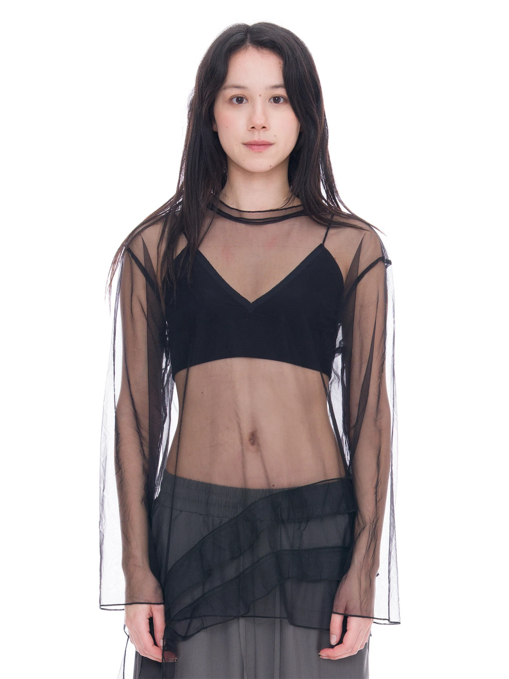 A model wearing a black sheer mesh long-sleeve crop top with a choker collar, layered over a black bralette, paired with a dark gray tiered skirt, against a solid white background.