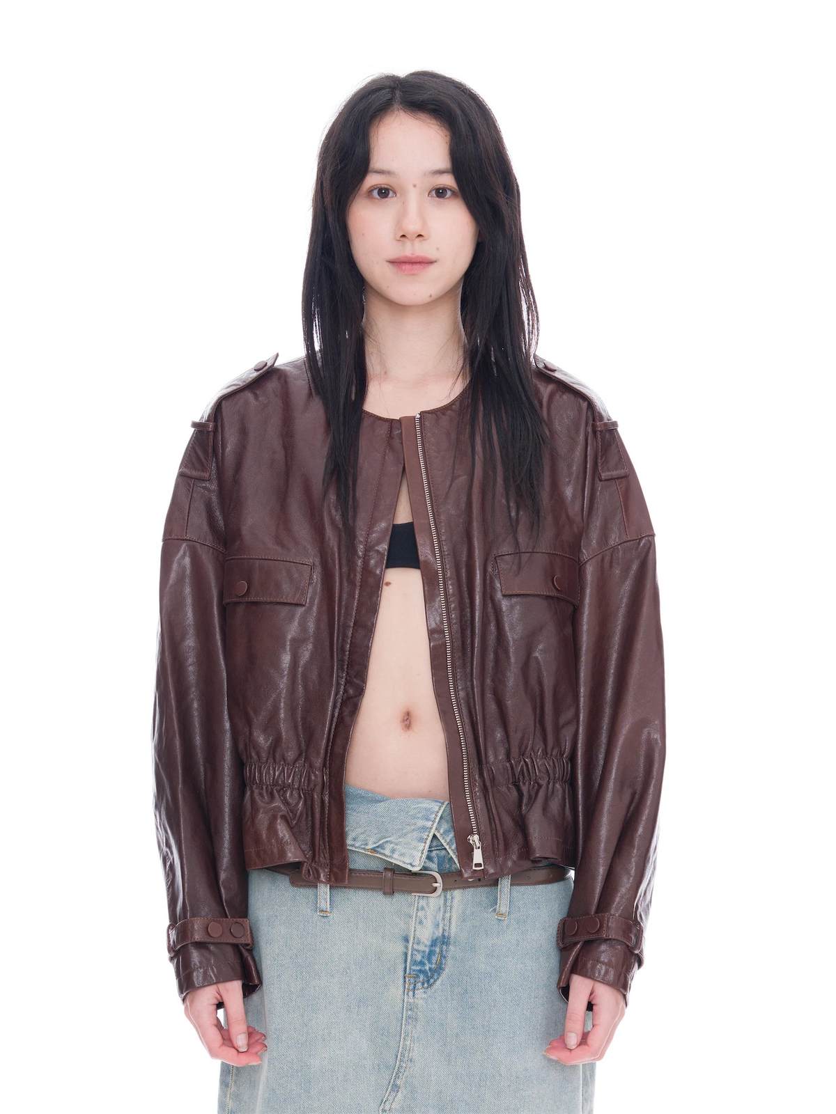 A model wearing a dark brown leather jacket with a front zipper, buttoned flap pockets, and elasticized cuffs, styled with light-wash denim jeans against a solid white background.
