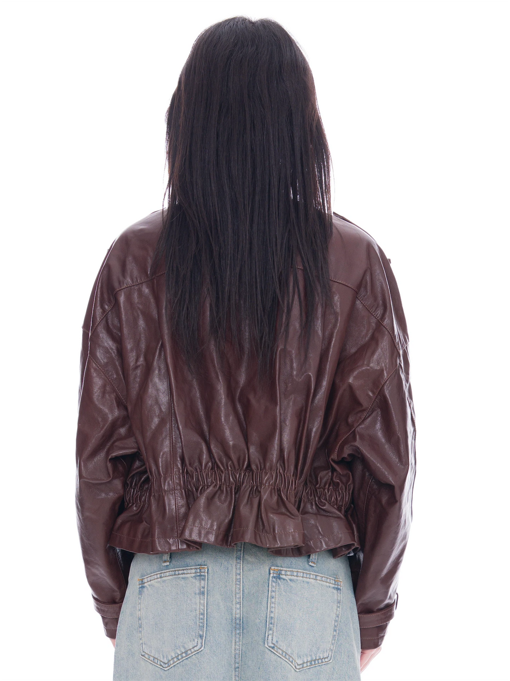 Rear view of a model with long dark hair wearing a brown sheep skin jacket with an elasticated back hem and light blue denim jeans, set against a plain white background.