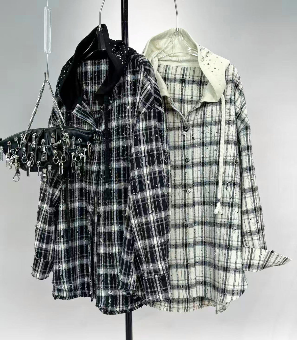 Two distressed plaid button-up shirts hanging on a black clothing rack, one in black and white plaid with a black studded collar, and the other in cream and white plaid with a cream studded collar and drawstring hood. A black studded shoulder bag hangs from the left.